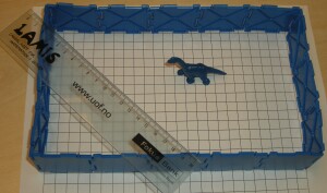 En linjal og en dinosaur gjerdet inn med jovo-brikker på et rutenett.